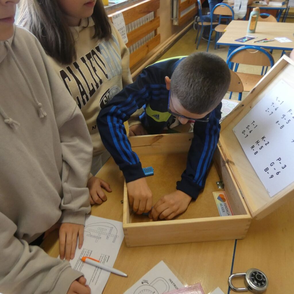 Uczniowie otwierają drewnianą skrzynkę z zagadkami podczas gry edukacyjnej
