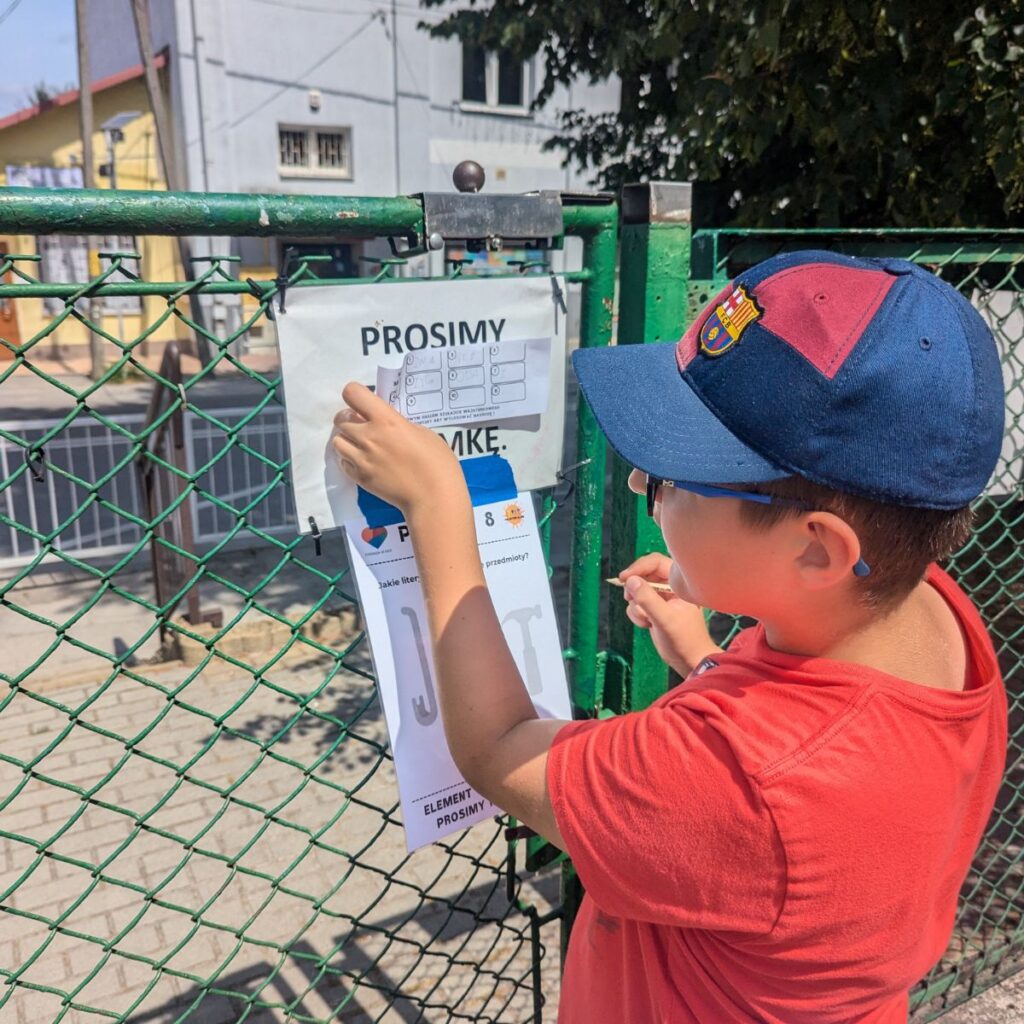 Dziecko przypina kartę z zadaniem do ogrodzenia w trakcie gry terenowej
