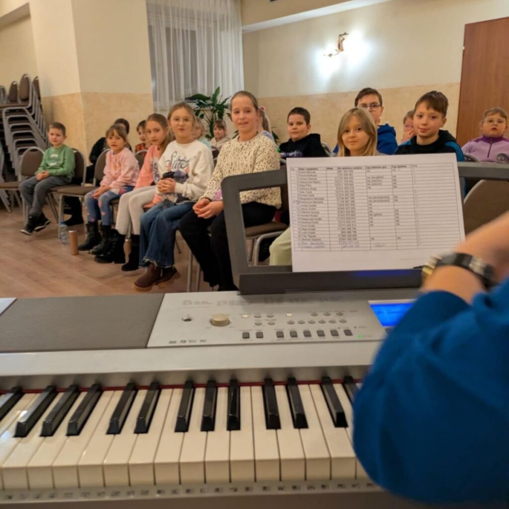Dzieci siedzące na krzesłach podczas zajęć muzycznych, w tle keyboard i nuty instruktora